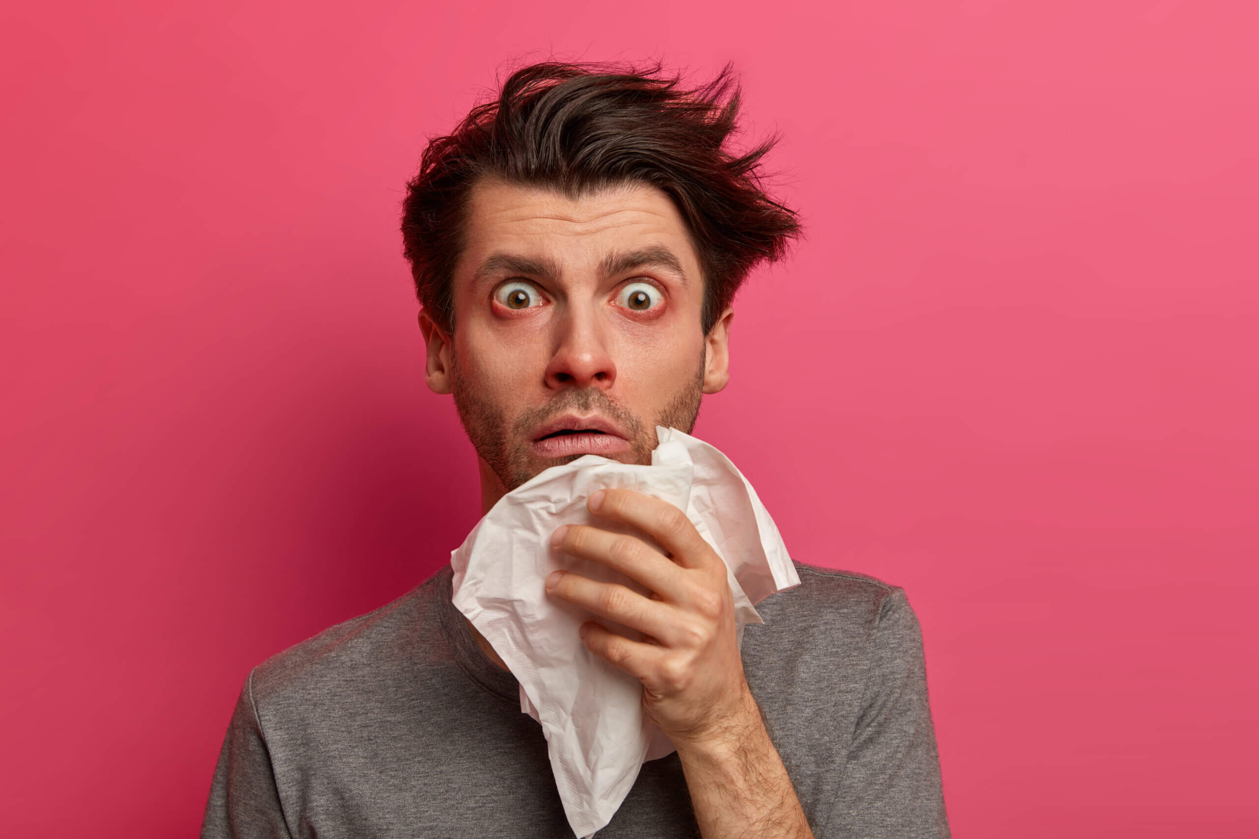 Stunned sick man has flu, virus or allergy respiratory, red watery eyes, blows nose in tissue, finds out about serious disease, poses over pink background. Health, medicine and symptoms concept